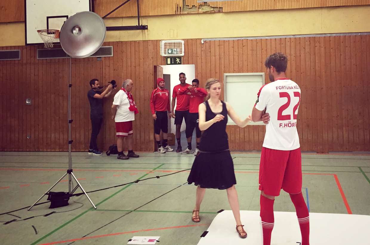 Eine Mitarbeiterin von mehrwert gibt einem Fortuna Köln Spieler Anweisungen für das Fotoshooting in einer Turnhalle