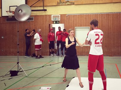 Eine Mitarbeiterin von mehrwert gibt einem Fortuna Köln Spieler Anweisungen für das Fotoshooting in einer Turnhalle